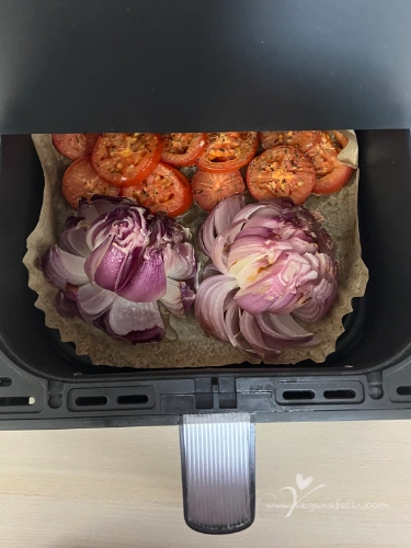 Cebolla cortada en forma de flor y rodajas de tomate en la cesta de una airfryer antes de cocinar.